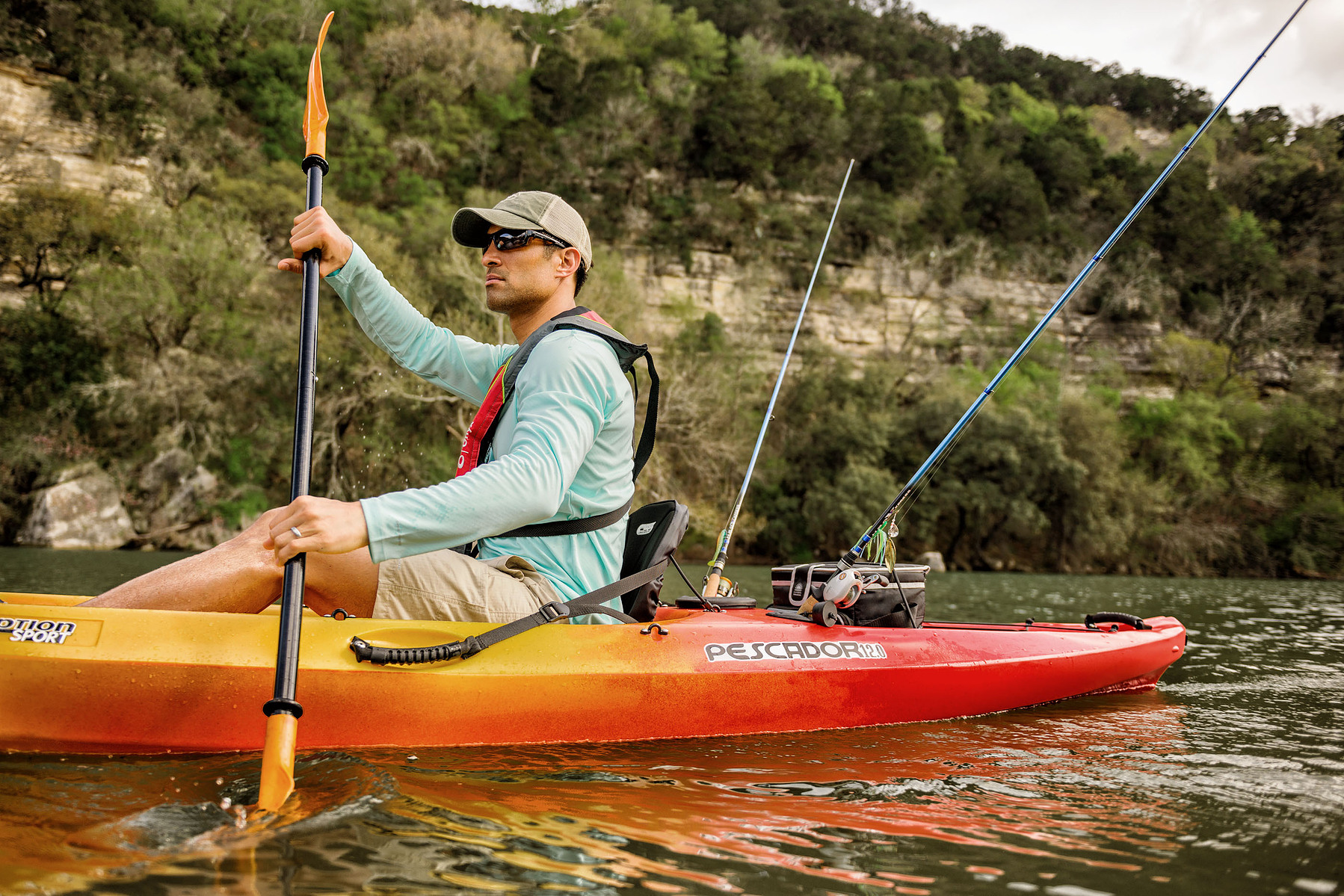 A person paddles an orange fishing kayak on a river with rods mounted behind the seat.
