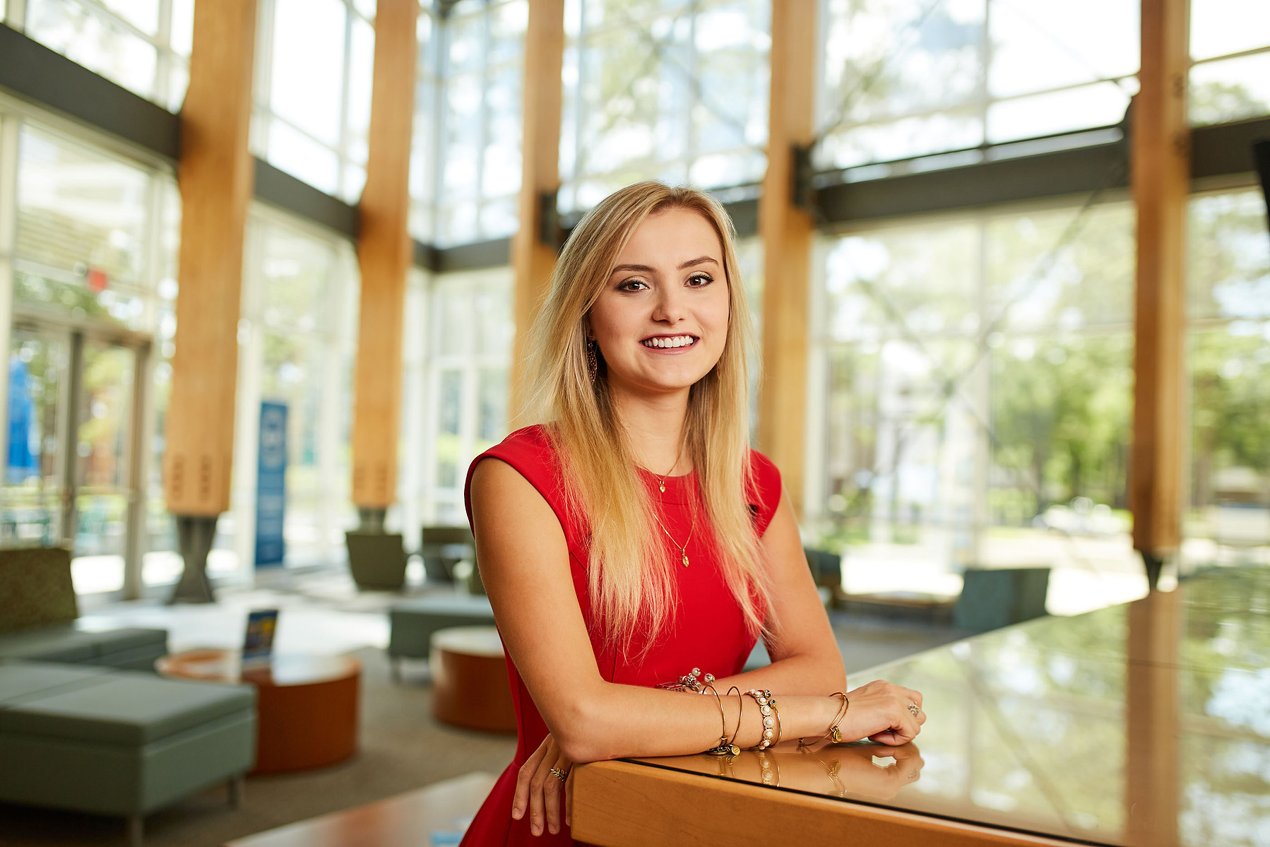 A smiling woman in a red sleeveless dress rests her hands on a table in a bright lobby.