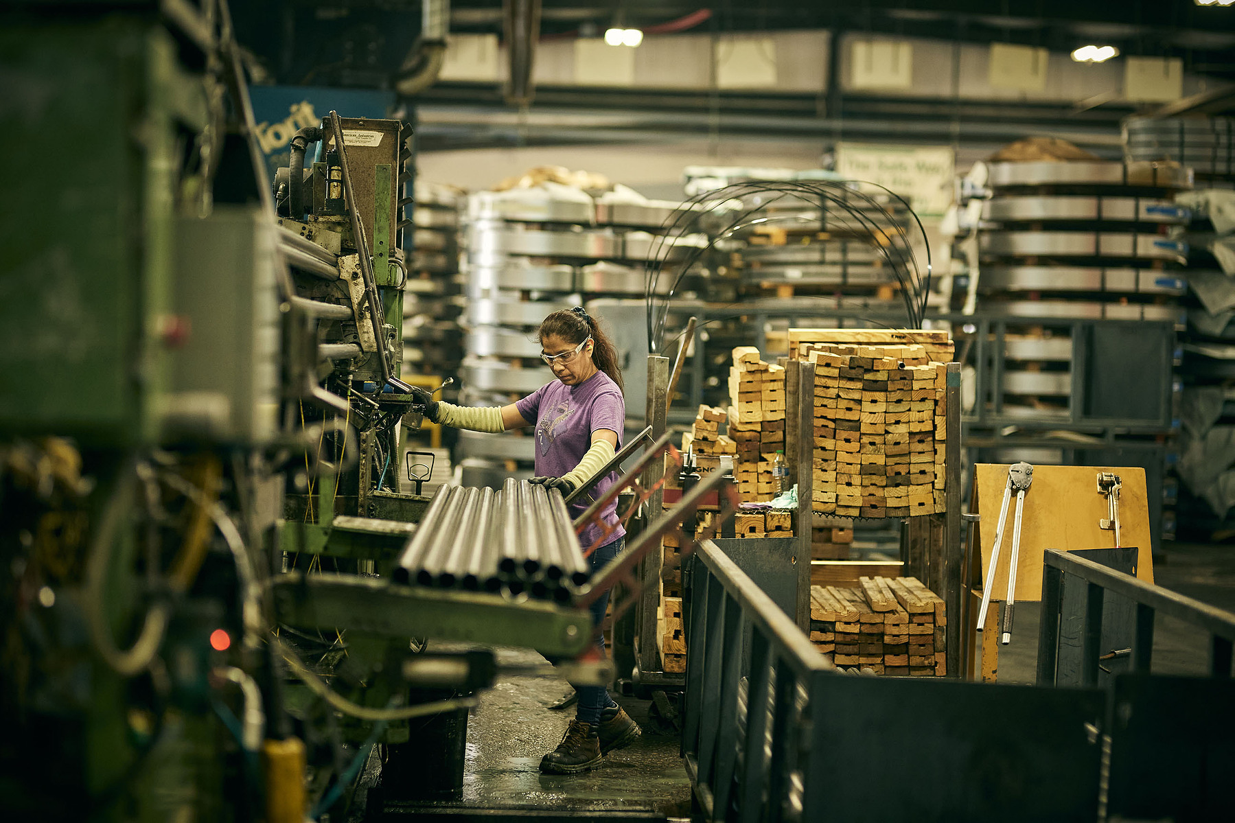 A worker operates equipment beside rows of metal parts on a factory production line.