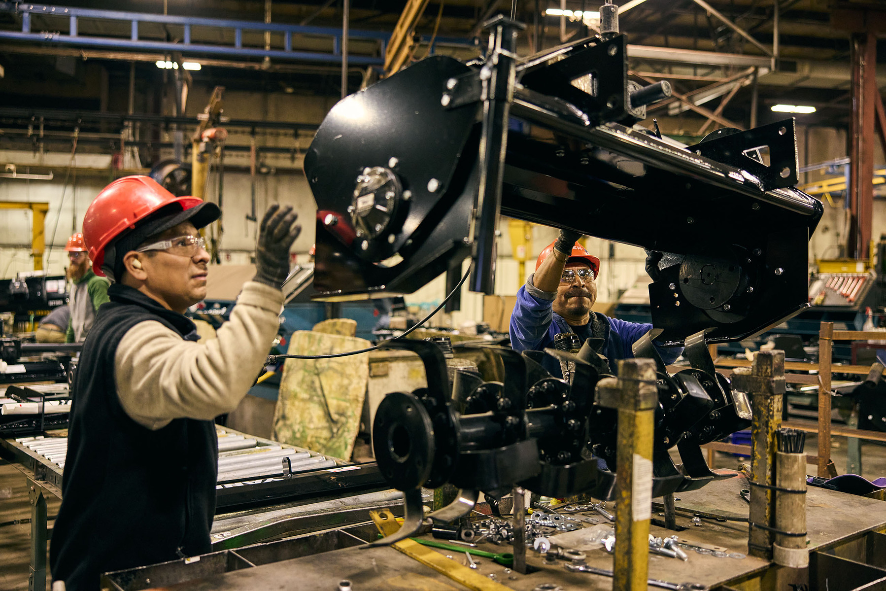 A worker in a red hard hat handles a black machine component suspended on an assembly line.