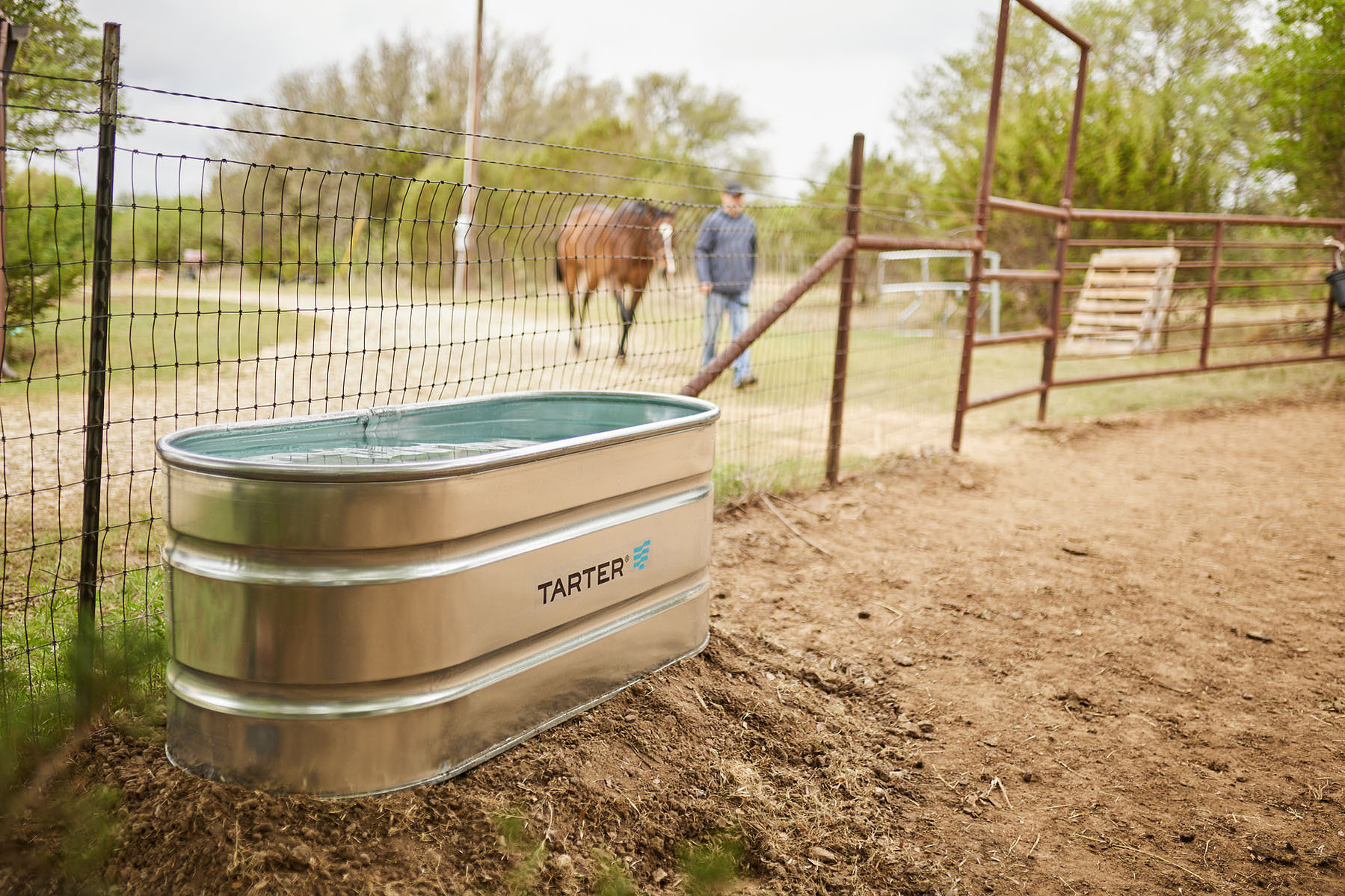 A galvanized stock tank sits beside a wire fence, with a horse and handler in the background.